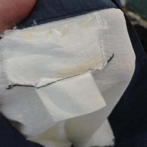 Puede incluir: Una etiqueta de prenda blanca con manchas amarillas visibles est&aacute; cosida en el cuello interior de una chaqueta azul marino oscuro. La etiqueta es rectangular con bordes deshilachados y est&aacute; unida con hilo negro. Una etiqueta de talla blanca m&aacute;s peque&ntilde;a est&aacute; unida debajo.