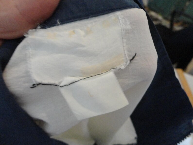 Puede incluir: Una etiqueta de prenda blanca con manchas amarillas visibles est&aacute; cosida en el cuello interior de una chaqueta azul marino oscuro. La etiqueta es rectangular con bordes deshilachados y est&aacute; unida con hilo negro. Una etiqueta de talla blanca m&aacute;s peque&ntilde;a est&aacute; unida debajo.