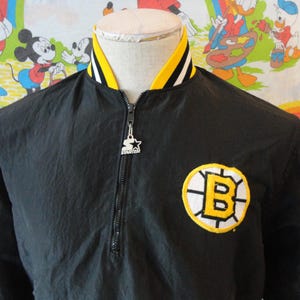 Puede incluir: Chaqueta negra Starter con cuello amarillo y blanco y cierre de media cremallera. La chaqueta presenta un parche con el logotipo de los Bruins de Boston en el pecho y un logotipo de Starter en el tirador de la cremallera.