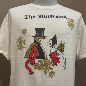 May include: White t-shirt with a black and white illustration of a man and woman in a winter scene. The text "The Nut Carol" is printed on the shirt.