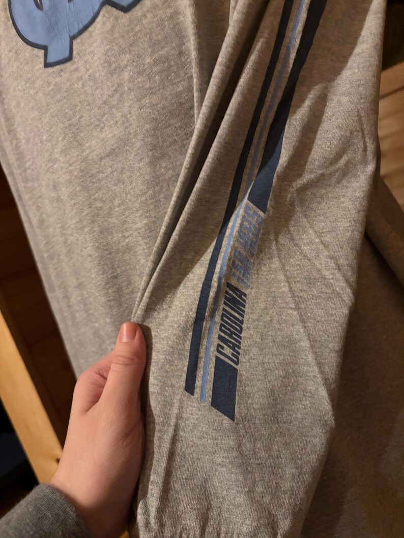 May include: Gray long-sleeve shirt with blue and white stripes on the sleeve. The text "CAROLINA" is printed on the sleeve.