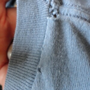 May include: Close-up of light blue fabric, possibly knit or jersey, with visible stitching and texture. The fabric appears soft and slightly worn, with subtle variations in color and a few small imperfections.