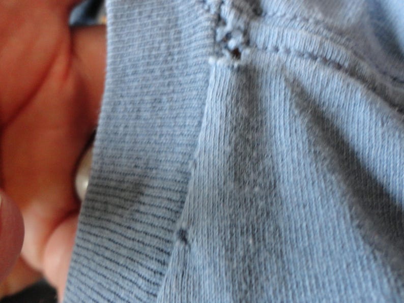 May include: Close-up of light blue fabric, possibly knit or jersey, with visible stitching and texture. The fabric appears soft and slightly worn, with subtle variations in color and a few small imperfections.