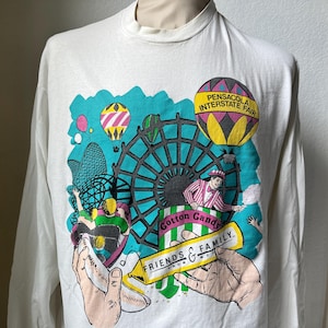 May include: White long-sleeved shirt with a fairground-themed graphic. The design includes a Ferris wheel, hot air balloons, and the text "Pensacola Interstate Fair." The shirt also has the words "Cotton Candy" and "Friends & Family."