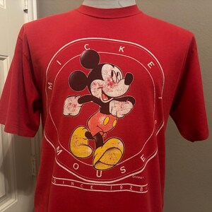 Op de afbeelding: Rood T-shirt met een witte cirkelvormige afbeelding met Mickey Mouse in een rood en geel outfit. De cirkelvormige afbeelding heeft de tekst "Mickey Mouse" en "Since 1928" rond de rand.