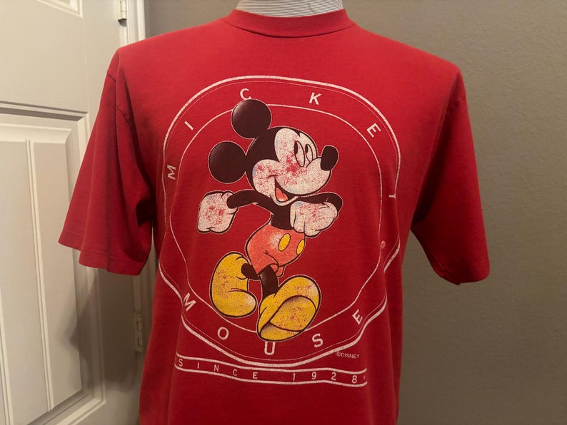 Op de afbeelding: Rood T-shirt met een witte cirkelvormige afbeelding met Mickey Mouse in een rood en geel outfit. De cirkelvormige afbeelding heeft de tekst "Mickey Mouse" en "Since 1928" rond de rand.