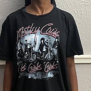 以下が含まれることがあります： Mötley Crüeのバンドロゴとグラフィックがプリントされた黒の半袖Tシャツ。ピンク色の文字でバンド名が記され、その下にはバイクに乗ったバンドメンバーのモノクロ写真と、ピンク色でGirls Girls Girlsというテキストがデザインされています。