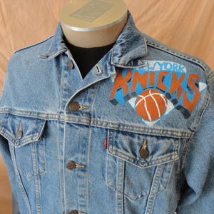 May include: Light blue denim jacket with the New York Knicks logo painted on the upper back. The logo features the team name in orange and blue, with a basketball in the center. The jacket has button closures and pockets.