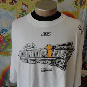 Op de afbeelding: Wit T-shirt met een zilveren en zwarte grafische afbeelding met de tekst "2005 NBA Finals The Finals 2005 Champions San Antonio Spurs", een basketbaltrofee en het logo van de San Antonio Spurs.