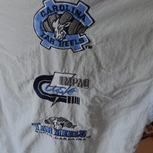 Puede incluir: Una chaqueta cortavientos blanca con logotipos bordados. El logotipo superior muestra una cabeza de carnero con el texto Carolina Tar Heels. El logotipo central muestra el texto IMPAQ USA. El logotipo inferior presenta una cabeza de carnero con el texto Tar Heels North Carolina.