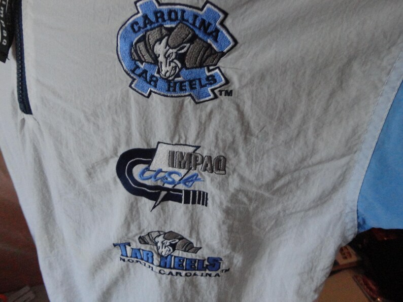 Puede incluir: Una chaqueta cortavientos blanca con logotipos bordados. El logotipo superior muestra una cabeza de carnero con el texto Carolina Tar Heels. El logotipo central muestra el texto IMPAQ USA. El logotipo inferior presenta una cabeza de carnero con el texto Tar Heels North Carolina.