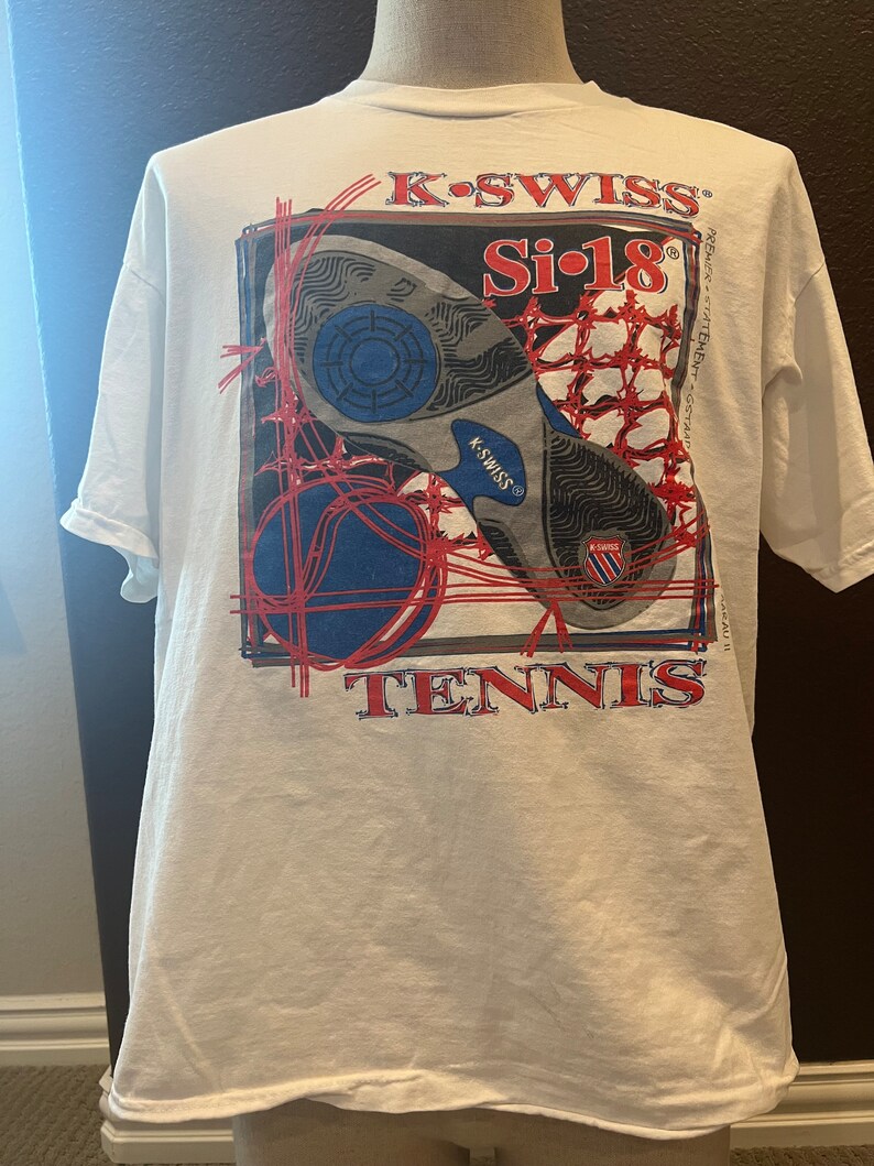May include: White t-shirt with a red, blue, and black graphic of a tennis shoe with the text "K-SWISS Si-18 TENNIS" on the back.