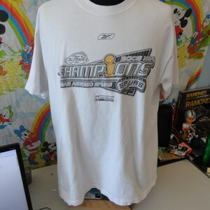May include: White t-shirt with the text "2005 NBA CHAMPIONS SAN ANTONIO SPURS" in black, silver, and gold. The Reebok logo is visible near the neckline. The shirt is displayed on a mannequin.