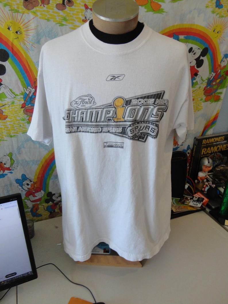 May include: White t-shirt with the text "2005 NBA CHAMPIONS SAN ANTONIO SPURS" in black, silver, and gold. The Reebok logo is visible near the neckline. The shirt is displayed on a mannequin.