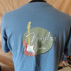 May include: A light blue t-shirt featuring the Fender logo with a red and white electric guitar graphic. The logo includes the text "Fine Electric Guitars Since 1946". The shirt is made of a soft material.