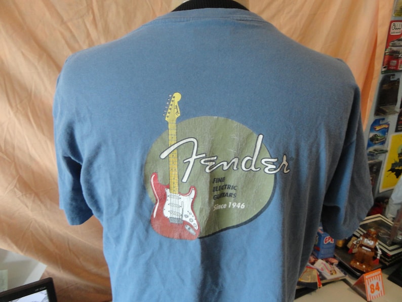 May include: A light blue t-shirt featuring the Fender logo with a red and white electric guitar graphic. The logo includes the text "Fine Electric Guitars Since 1946". The shirt is made of a soft material.