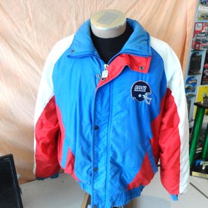 Puede incluir: Una chaqueta azul, roja y blanca con el logotipo de los Giants en el pecho. La chaqueta tiene cierre de cremallera y botones de presión. El cuello es azul y las mangas son blancas. La chaqueta se exhibe en un maniquí.