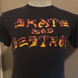 May include: Black t-shirt with the words "SKATE AND DESTROY" in a stylized, flame-like font. The text is in shades of yellow, orange, and red, creating a fiery effect. The shirt is short-sleeved.