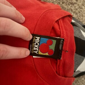 Op de afbeelding: Rood T-shirt met een Mickey Mouse Unlimited Disney-label. Het label is zwart met een gele, groene, blauwe en rode achtergrond. Het Mickey Mouse-hoofd is rood met zwarte contouren.