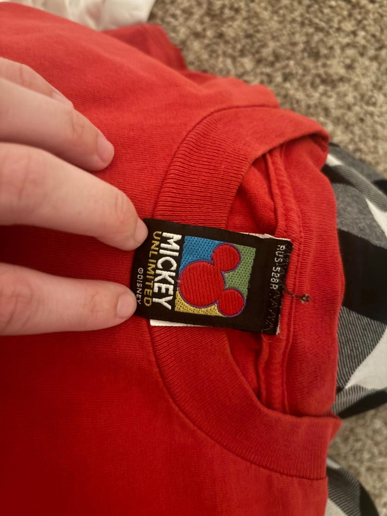 Op de afbeelding: Rood T-shirt met een Mickey Mouse Unlimited Disney-label. Het label is zwart met een gele, groene, blauwe en rode achtergrond. Het Mickey Mouse-hoofd is rood met zwarte contouren.