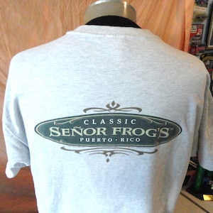 May include: Light gray t-shirt with the text "CLASSIC SEÑOR FROG'S PUERTO RICO" in a dark green and brown oval design. The shirt is displayed on a mannequin. The background is a light peach color.