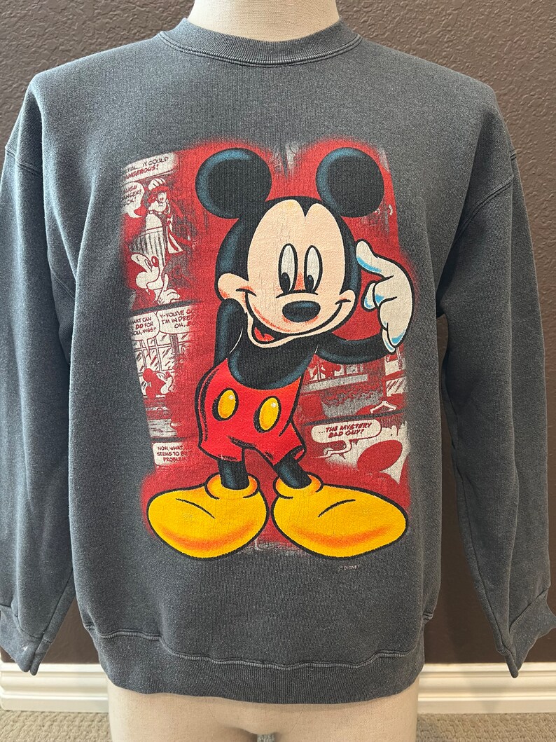 May include: A gray sweatshirt with a vintage Mickey Mouse graphic. The graphic features Mickey Mouse in a red and yellow outfit, with a red background and comic book style text. The text reads "The Mystery Bad Guy!"