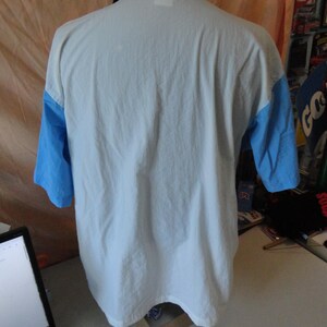Puede incluir: Un polo de manga corta con cuerpo blanco, mangas azul claro y cuello azul marino oscuro. La prenda se muestra desde la parte posterior, revelando el panel trasero blanco liso, las mangas en contraste y el cuello doblado de color marino.
