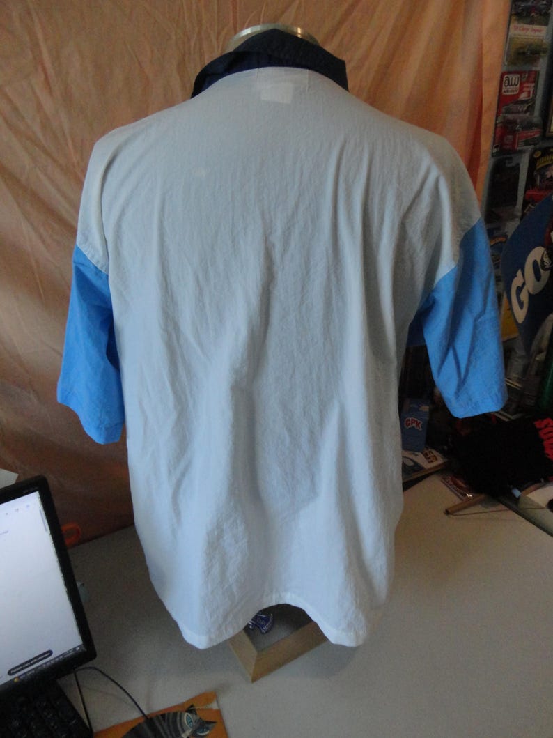Puede incluir: Un polo de manga corta con cuerpo blanco, mangas azul claro y cuello azul marino oscuro. La prenda se muestra desde la parte posterior, revelando el panel trasero blanco liso, las mangas en contraste y el cuello doblado de color marino.