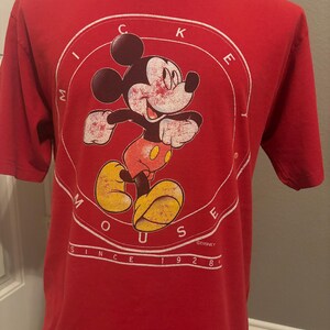 Op de afbeelding: Rood T-shirt met een vintage Mickey Mouse-afbeelding. De afbeelding toont Mickey Mouse in een cirkel met de woorden "Mickey Mouse" eromheen. De tekst "Since 1928" staat onder de cirkel. &copy;DISNEY