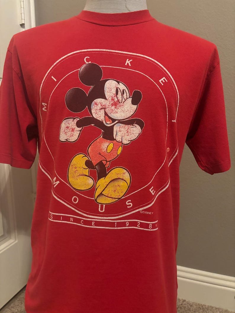 Op de afbeelding: Rood T-shirt met een vintage Mickey Mouse-afbeelding. De afbeelding toont Mickey Mouse in een cirkel met de woorden "Mickey Mouse" eromheen. De tekst "Since 1928" staat onder de cirkel. &copy;DISNEY