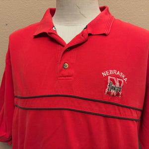 May include: Red polo shirt with a black and white embroidered Nebraska Huskers logo. The shirt has two black stripes across the chest.