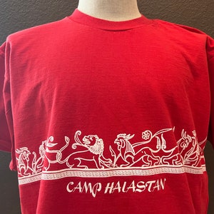Puede incluir: Camiseta roja con un diseño blanco de un león, un pájaro y otras criaturas míticas. El diseño incluye el texto "CAMP HALASTAN".