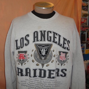 May include: A light gray crewneck sweatshirt featuring the text LOS ANGELES RAIDERS in black. The design includes a Raiders shield logo, an AFC West emblem, and an NFL logo. Printed details include team colors, stadium address, and training camp location.