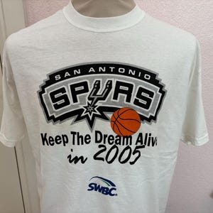 Op de afbeelding: Wit T-shirt met het San Antonio Spurs logo en een basketbalafbeelding. Het shirt heeft de tekst "Keep The Dream Alive in 2005" en het SWBC-logo.