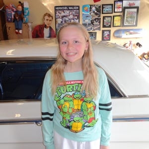 Könnte beinhalten: Eine Person trägt ein hellblaues Langarmshirt mit den Teenage Mutant Ninja Turtles. Das Shirt hat schwarze Streifen an den Ärmeln. Die Person steht vor einem weißen Auto.