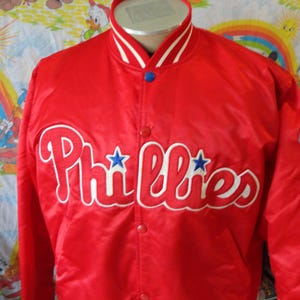 Peut inclure: Veste rouge en satin avec le mot "Phillies" en blanc sur le devant. La veste a un col et des poignets blancs et bleus, avec des étoiles bleues. Un écusson du logo de l'équipe est sur la manche droite.
