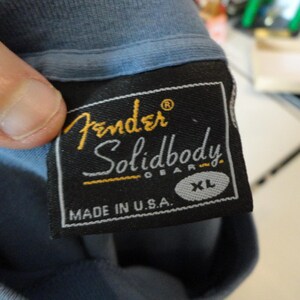 May include: Close-up of a black Fender Solidbody Gear XL label sewn onto a blue t-shirt. The label features the Fender logo in gold and the text "Solidbody Gear" in white. The words "Made in U.S.A." are also visible.