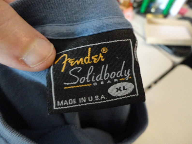 May include: Close-up of a black Fender Solidbody Gear XL label sewn onto a blue t-shirt. The label features the Fender logo in gold and the text "Solidbody Gear" in white. The words "Made in U.S.A." are also visible.