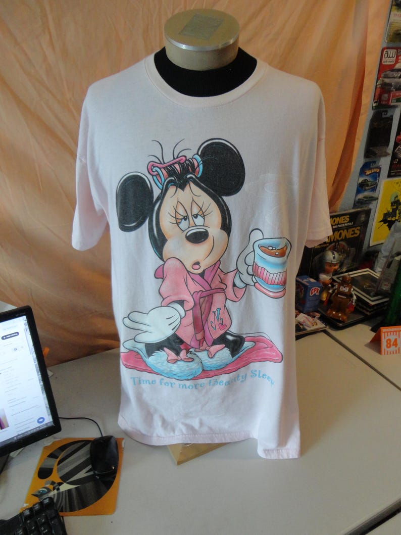 May include: A light pink t-shirt featuring a cartoon Minnie Mouse in a pink robe, holding a cup. The text "Time for more Beauty Sleep" is below the image. The shirt is displayed on a mannequin.