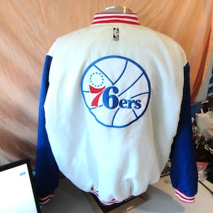 May include: A white and blue varsity jacket featuring the Philadelphia 76ers logo on the back. The jacket has a white body, blue sleeves, and a ribbed collar with red and white stripes. The NBA logo is embroidered near the collar.