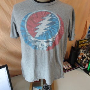 May include: A gray t-shirt featuring a colorful graphic of a skull with a lightning bolt. The graphic is in a circle with red, blue, and white tie-dye patterns. The shirt is displayed on a mannequin.