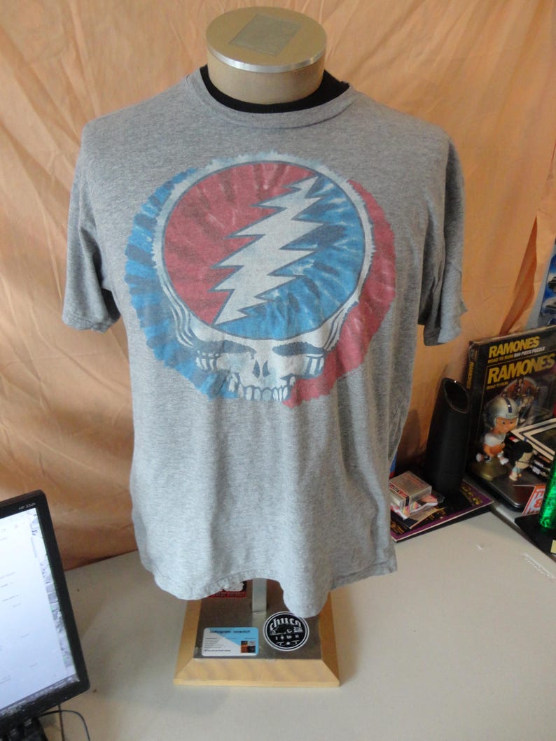 May include: A gray t-shirt featuring a colorful graphic of a skull with a lightning bolt. The graphic is in a circle with red, blue, and white tie-dye patterns. The shirt is displayed on a mannequin.