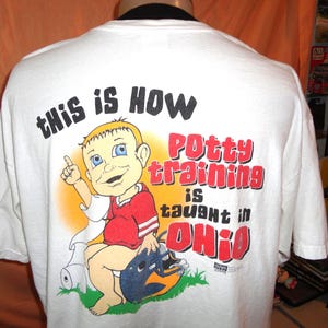May include: White t-shirt featuring a cartoon illustration of a child on a potty, with the text "THIS IS HOW Potty training is taught in OHIO". The child wears a red shirt and a blue helmet.