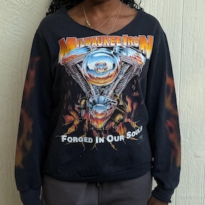 May include: Black long-sleeve sweatshirt with a motorcycle engine graphic and the text "Milwaukee Iron Forged in Our Souls." The sleeves have a flame design. The sweatshirt has a v-neck.