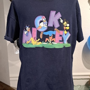 May include: A navy blue t-shirt with a graphic print of Mickey Mouse, Donald Duck, Goofy, and Pluto. The characters are arranged around the word "Mickey" in purple letters.