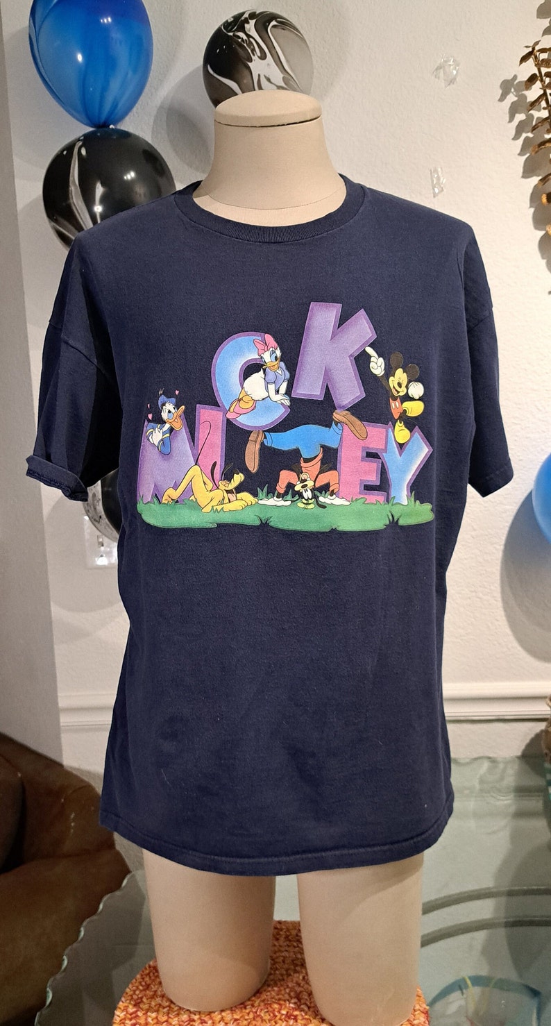 May include: A navy blue t-shirt with a graphic print of Mickey Mouse, Donald Duck, Goofy, and Pluto. The characters are arranged around the word "Mickey" in purple letters.