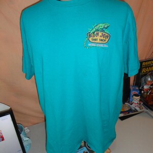 May include: Teal short-sleeved t-shirt featuring a green iguana graphic and the text "Ron Jon Surf Shop, Cocoa Beach, FL". The shirt is displayed on a mannequin.