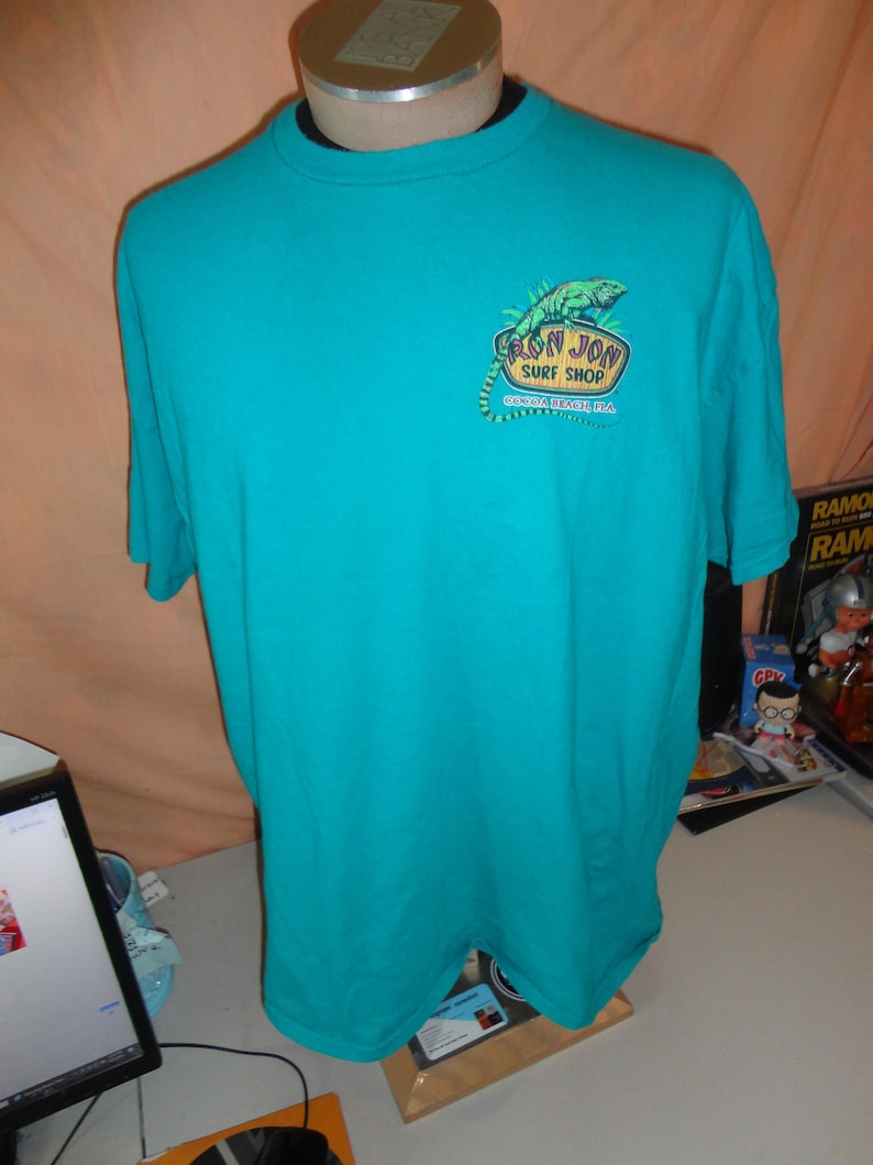 May include: Teal short-sleeved t-shirt featuring a green iguana graphic and the text "Ron Jon Surf Shop, Cocoa Beach, FL". The shirt is displayed on a mannequin.