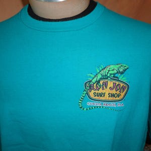 May include: Teal t-shirt with a black collar. The shirt displays a graphic of a green iguana and the words "RON JON SURF SHOP COCOA BEACH, FLA" in a logo design.
