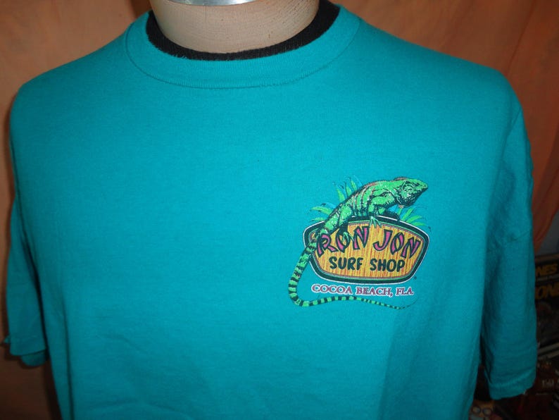 May include: Teal t-shirt with a black collar. The shirt displays a graphic of a green iguana and the words "RON JON SURF SHOP COCOA BEACH, FLA" in a logo design.